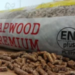 pellet lapwood w worku bokiem, rozsypany pellet obok, biały worek z żółtym i czerwonym elementem płomienia